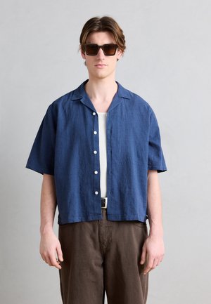 Junger Mann mit dunkler Sonnenbrille, blauem kurzärmligem Hemd mit Knöpfen, weißem Unterhemd und brauner Hose vor hellgrauem Hintergrund.