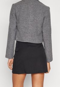 Osoba v šedém křížovém saku s krátkým rukávem a černé sukni je zobrazena zezadu, což ukazuje na detaily outfitu.