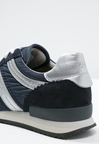 Mörkblå sportskor med en blank silverklack, texturerade mockadetaljer och vitt randigt mönster. Gummisula för bra grepp.