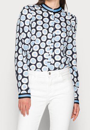 Nainen, joka käyttää pitkähihainen napillista puseroa, jossa on sinivalkoinen pyöreä kuvio, tunnettuna valkoisiin, korkeavyötäröisiin farkkuihin.