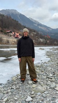 Ένα άτομο στέκεται σε μια βραχώδη ακτή δίπλα σε ένα ποτάμι, φορώντας μαύρο μπουφάν με κουκούλα, χακί παντελόνι και καφέ μπότες με μαύρες σόλες.