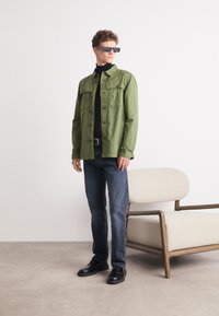 Zelená bavlněná bunda s náprsními kapsami, černý rolák, tmavě modré džínsy a černé šněrovací boty. Model stojí vedle béžové židle.