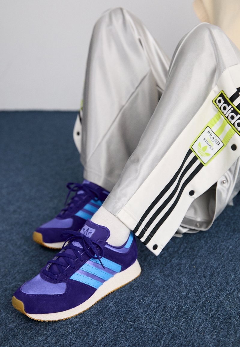 Personne portant un pantalon de survêtement Adidas argenté avec des bandes noires et des baskets Adidas violettes, assise sur un sol tapissé de moquette bleue.