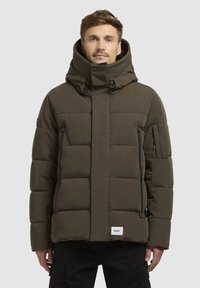 Smešani smeđi kaput s visokim ovratnikom, višestrukim džepovima na patentni zatvarač i pravokutnom oznakom pri rubu. Teksturirana tkanina.