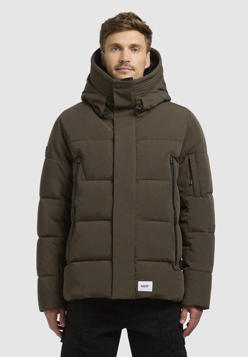 Braune gepolsterte Jacke mit hohem Kragen, mehreren Reißverschluss-Taschen und einem rechteckigen Logo-Patch am Saum. Strukturierte Stoffoberfläche.