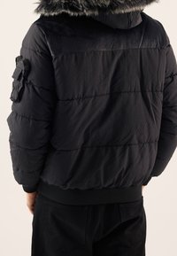 Czarna kurtka puchowa z kapturem wykończonym futerkiem, posiadająca poziomą pikowaną fakturę, ściągacze na rękawach oraz kieszeń na lewym rękawie.