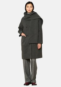 Cappotto grigio in lana con un ampio colletto drappeggiato, doppi bottoni, tasche frontali e una vestibilità comoda, abbinato a pantaloni argento strutturati e scarpe bordeaux.