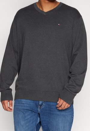 Mann trägt einen dunkelgrauen V-Ausschnitt-Pullover mit kleinem Logo auf der Brust und blaue Jeans, steht vor einem einfarbigen Hintergrund.