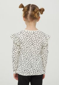 Koszula z długim rękawem w kremowym kolorze z czarnym kropkowanym wzorem, z falbankami na ramionach i okrągłym dekoltem, noszona z czarnymi spodniami.