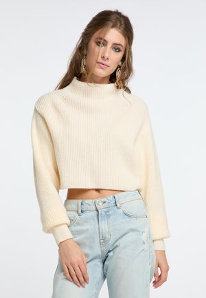 Pull court crème à côtes, avec un col haut et des manches larges, associé à un jean léger bleu délavé. Texture lisse, design décontracté.