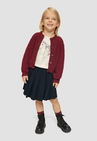 Bordeaux gebreide cardigan over een wit shirt met een grafisch ontwerp, gecombineerd met een geplooide marineblauwe rok en zwarte veterschoenen.