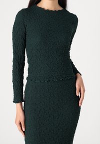 Haut en maille texturée verte à manches longues avec un col festonné, associé à une jupe ajustée assortie. Présente un design en tissu froissé.