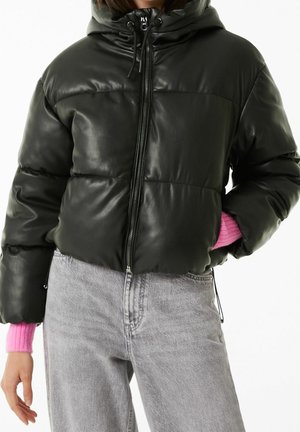 Black padded jacket with hood and zipper worn over a pink sweater, paired with light gray high-waisted jeans, hand in pocket.
