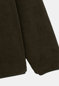 Veste en polaire marron avec une surface texturée, des bords arrondis et une coupe légèrement décontractée, avec un bord noir le long de l'ourlet.