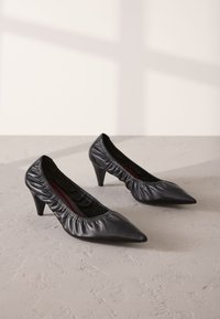 Black leather pointed-toe pumps, featuring a gathered upper design and a mid-height stiletto heel, with a smooth texture and subtle sheen.