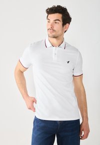 Valge polo särk punase ääremiseks kaeluses ja varruka servades, vasakul rinnal väike must logo. Sile kangas, lühikesed varrukad.