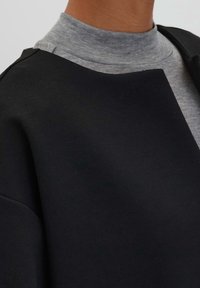 Svart, texturerad ytterlager med en hög, rundad halsringning som matchar en grå, åtsittande polotröja under.