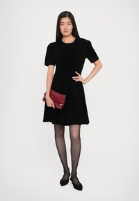 Woman wearing a black short-sleeve dress, black tights, black shoes, holding a red clutch purse, standing against a plain light background.