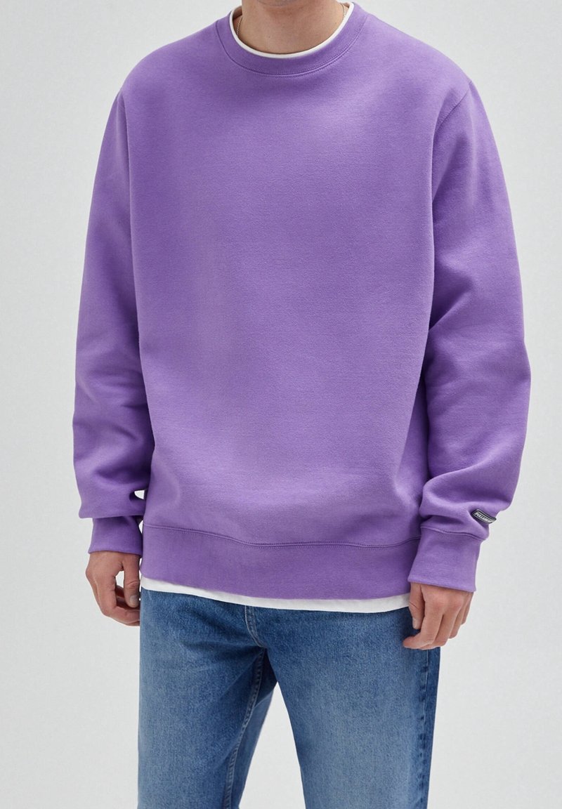 Sweatshirt violet à col rond en tissu doux, avec une coupe décontractée et des poignets côtelés. Porté par-dessus une chemise blanche, associé à un jean en denim.