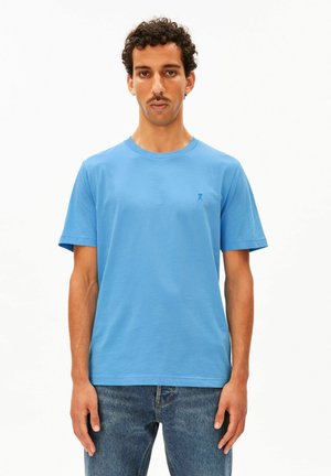 Homme aux cheveux bouclés et à la moustache, portant un t-shirt bleu clair uni à manches courtes et un jean bleu sur fond blanc.