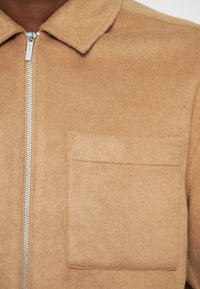 Tan wool blend shirt jacket featuring a zipper closure, pointed collar, and a single chest pocket. Soft texture with a clean design.