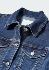 Veste en denim bleu avec des boutons en métal, des coutures surpiquées et deux poches poitrine, montrant un col plié et une étiquette de marque à l'intérieur.