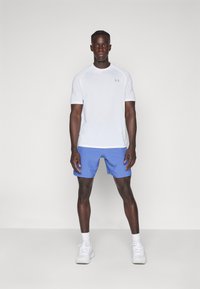 Weißes, kurzärmeliges Sportshirt mit einem hellgrauen Logo, kombiniert mit blauen Shorts. Das Model trägt weiße Sneakers und zeigt eine neutrale Haltung.