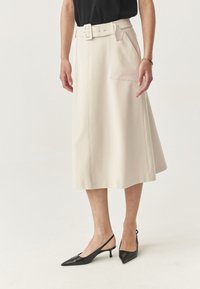 Donna che indossa una gonna midi beige con una cintura, un top nero, scarpe slingback con tacchi kitten neri e un braccialetto d'oro sul polso destro.