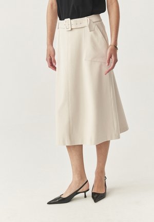 Frau in einem beigefarbenen Midikleid mit einem Gürtel, einem schwarzen Oberteil, schwarzen Slingback-Pumps mit Kitten-Heels und einem goldenen Armband am rechten Handgelenk.