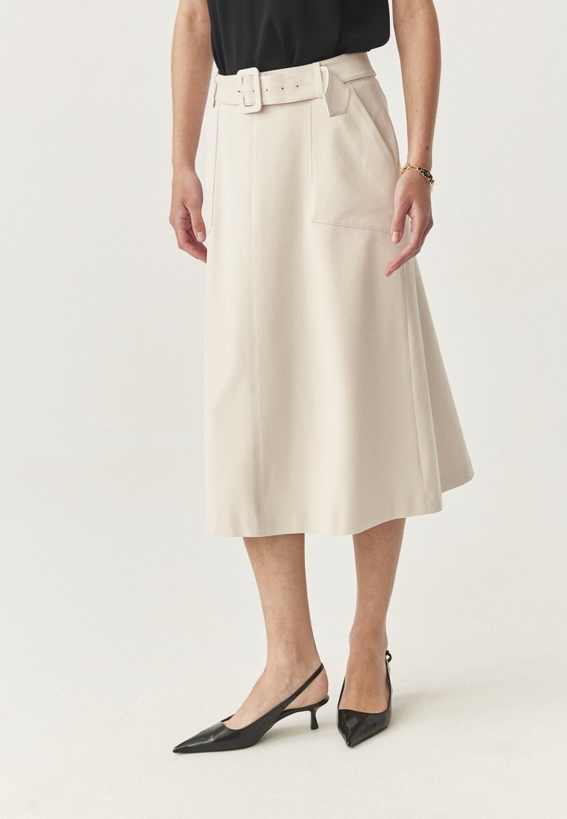 Donna che indossa una gonna midi beige con una cintura, un top nero, scarpe slingback con tacchi kitten neri e un braccialetto d'oro sul polso destro.