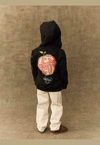Svart hettegenser med fargerik eplegrafikk på baksiden, med teksten "LES", beige bukser og brune sko.