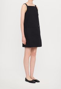 Vestido negro sin mangas con escote cuadrado, corte recto y detalle de cinturón en la cintura. Combinado con zapatos planos negros.