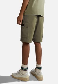 Pantaloni cargo verde oliva con tasche laterali, una vestibilità rilassata e una texture leggermente ruvida. Indossati con sneakers bianche e calze a righe.