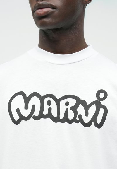 Detailní záběr osoby v bílé košili s černým, tučným logem v bublinkovém stylu s nápisem "Marni" na hrudi.