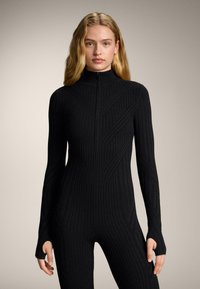 Žena s dugom plavom kosom u uskom crnom rebrastom kombinezonu s otvorima za palce i prednjim patentnim zatvaračem, stoji na neutralnoj pozadini.