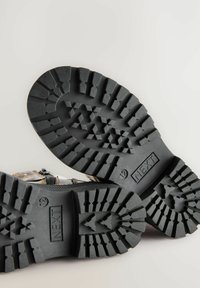Schwarze Gummisohle mit tiefen Profilmustern. Enthält ein Markenlabel "NEXT" und die Größe "12". Das Oberdesign besteht aus einer Mischung von Materialien und Farben.