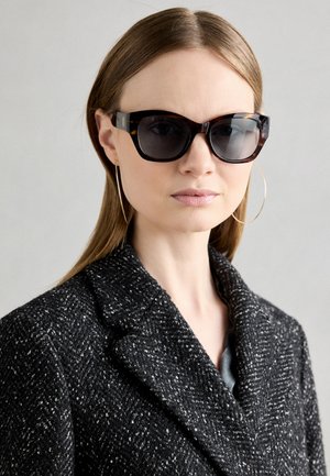 Schildpatt-Sonnenbrille mit dunklen Gläsern, breiten Rahmen und goldenen Akzenten. Getragen mit einem strukturierten schwarz-weißen Fischgrätenmantel.