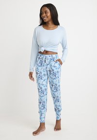 Maglietta azzurra a maniche lunghe con nodo frontale, abbinata a pantaloni jogger blu a fantasia con design floreale e polsini elastici.