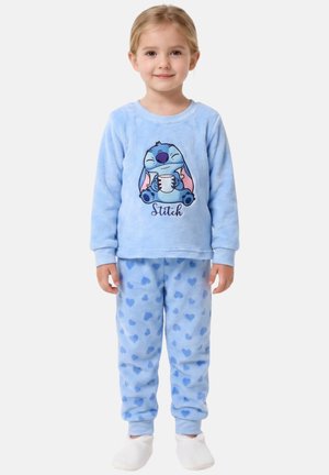 Jeune fille en pyjama polaire bleu avec le personnage Stitch tenant une tasse sur le haut et des motifs en forme de cœur sur le pantalon, portant des chaussons blancs.