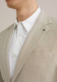 Beige Blazer mit dezenten hellbraunen Karos, strukturiertem Stoff und Notch-Kragen; kombiniert mit einem weißen Hemd darunter.