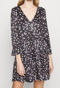 Robe noire à fleurs avec des manches longues, décolleté en V et jupe à volants. Présente des motifs floraux colorés sur un fond à pois. Tissu léger.