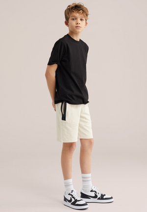 Boy with curly hair stands wearing black t-shirt, cream shorts with zipper detail, white striped socks, and black-and-white Nike sneakers.