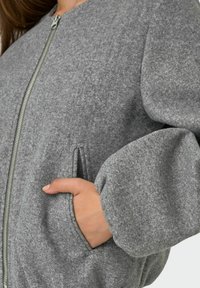 Sivi bombarder jakna od flisa s prednjim zatvaračem, bočnim džepovima i voluminoznim rukavima za opušteni izgled. Mekana tekstura i minimalistički dizajn.