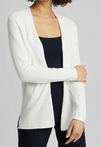 Bílý kardigan vyrobený z žebrované tkaniny, s otevřeným předním dílem, dlouhými rukávy, volným střihem a bočními rozparky pro snadnější pohyb.