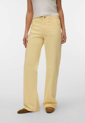 Moteris dėvi šviesiai geltonas, aukšto liemens, platėjusias platėjančias džinsines kelnes, derina jas su rudos spalvos lygiais batais ir balta rankovių neturinčia palaidine, stovi prieš baltą foną.