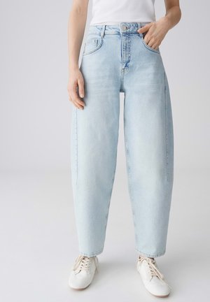 Persona che indossa jeans a vita alta di colore azzurro chiaro a gamba larga, sneakers bianche e una maglia bianca, con una mano in tasca e l'altra lungo il corpo.