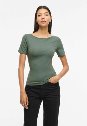 Kvinde med langt mørkt hår iført en tætsiddende grøn kortærmet top og sorte bukser med den ene hånd i lommen mod en ensartet hvid baggrund.