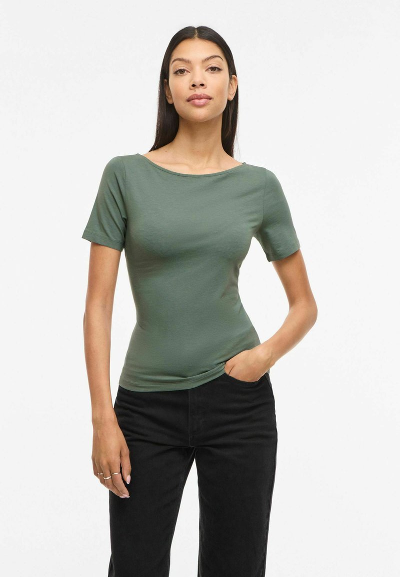 Hosszú sötét hajú nő testhezálló zöld rövid ujjú felsőt és fekete nadrágot visel, egyik kezét a zsebében tartja, egyszerű fehér háttér előtt.