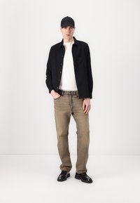 Black button-up shirt over a white T-shirt, light brown jeans, and black shoes. Accessorized with a black cap and silver earrings.