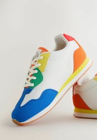 Valkoinen tennari, jossa sinisiä, vihreitä, keltaisia, oransseja ja punaisia korostuksia, valkoiset nauhat ja purukumia muistuttava pohja, vaaleaa taustaa vasten.